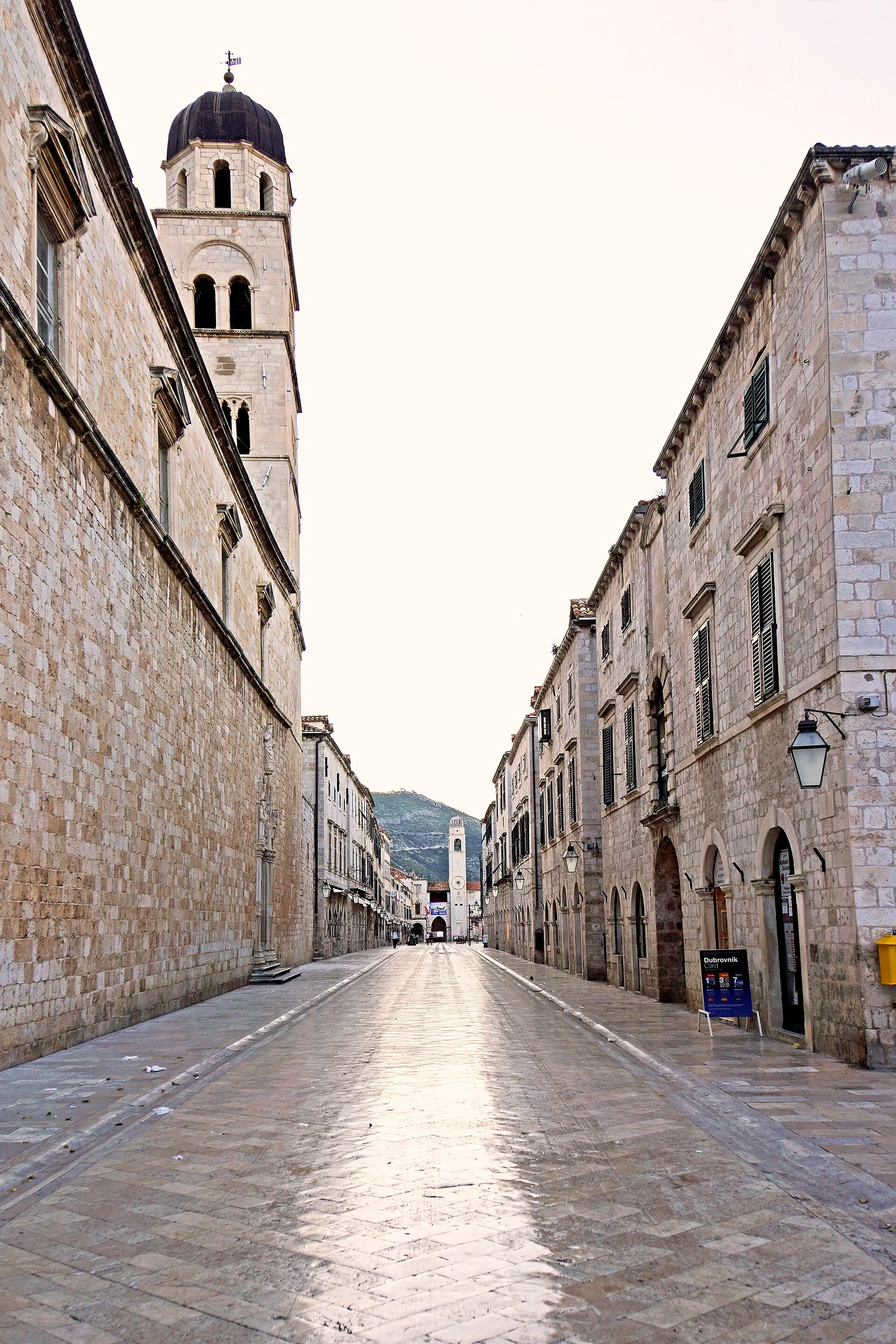 Der menschenleere Stradun in Dubrovnik am frühen Morgen mit poliertem Kalksteinpflaster, Franziskanerkirchturm links und Glockenturm am Ende der Gasse.