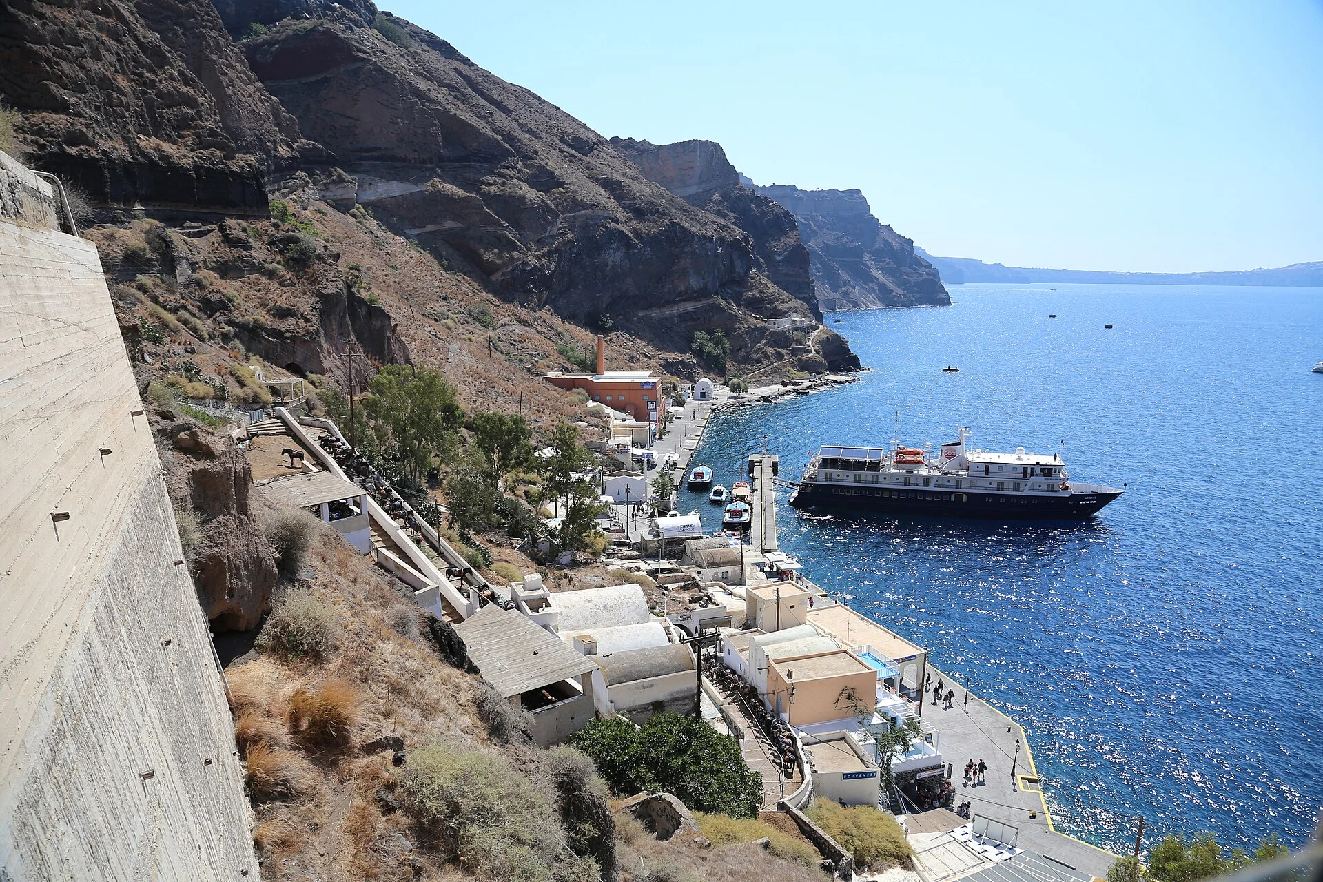 Der alte Hafen Skala unterhalb von Fira auf Santorin mit Tenderschiff, Eselspfad und Seilbahntrasse an der Calderawand.